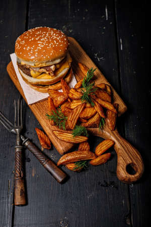 Fried potato wedges with herbs and delicious double burger on a wooden cutting board. Rustic dark...