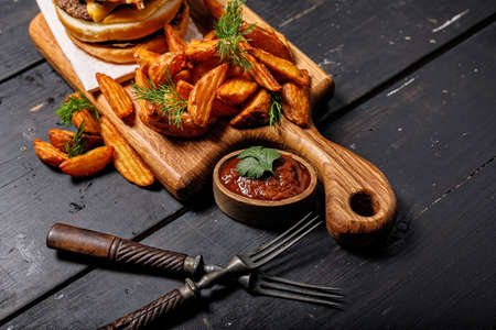 Appetizing fried potato wedges and burger on a wooden cutting board. Big portion of baked potatoes...