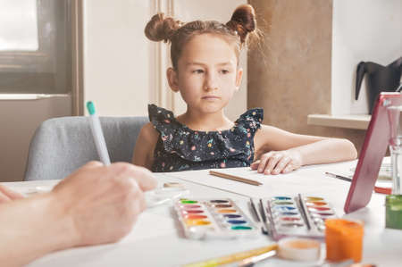 A cute beautiful girl is sitting at a table and watching an online drawing lesson. The child is at home due to the COVID-19 quarantine Online and internet technologies in everyday life. Lifestyleの写真素材