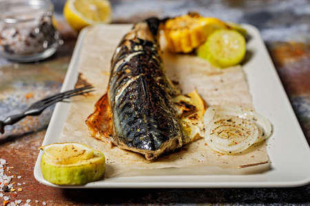 Close-up delicious roasted mackerel with vegetables on a rectangular plate. Appetizing grilled sea fish with corn, zucchini and spiced lemon. Mediterranean Kitchen. Healthy seafood. Spices and forks.の写真素材