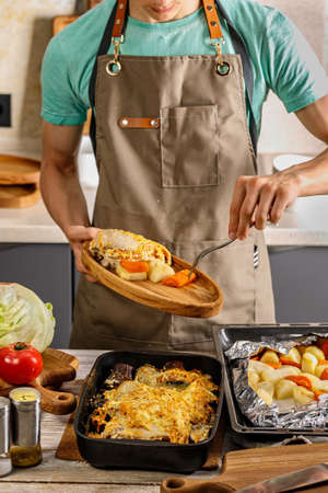 A young chef in a brown apron lays food on a wooden plate. On the table are baked chicken breast and vegetables for a dinner. Cooking tasty and healthy food in your home kitchen. Vertical shotの写真素材