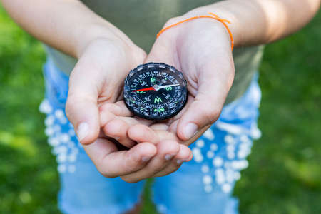 Girl holding the compass. Hands of a teenager holding a liquid compass. Red compass needle points north. Green grass background. Copy space. Orienteering on the ground. Selecting the directionの写真素材