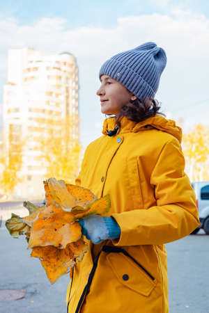 A smiling cute girl in a bright jacket with an autumn bouquet of bright fallen leaves. Portrait of a happy positive girl. Sunny fall day background. Side view. Vertical shot. Autumn moodの写真素材