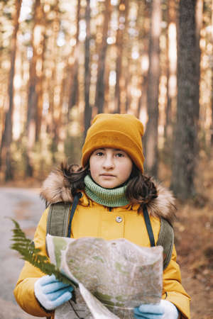 Cute caucasian girl is hiking in the autumn pine forest. A teenager with a backpack and a map walks along the route. Outdoor activity. Vertical shotの写真素材