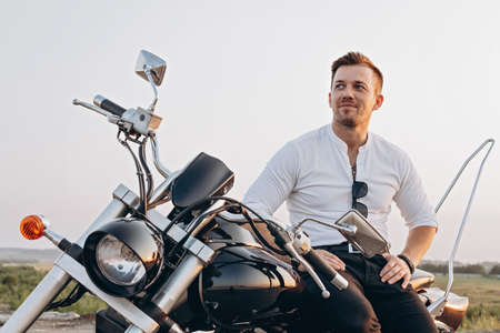 Stylish young attractive guy is sitting on a motorcycle. A nice young man in a white shirt sits and rests on a bike. Hot sunny day. Sky backgroundの写真素材