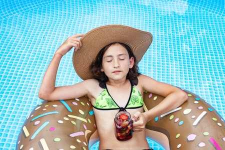 Beautiful girl in the blue swimming pool. Cute pretty teenager in a straw hat with inflatable donut and a berry cocktail. Teenager stock photo. Summer vacation concept.の写真素材