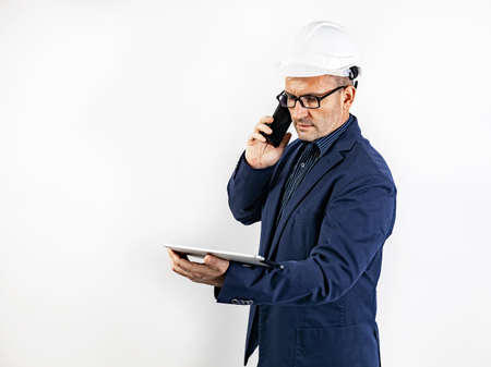 A male engineer in a white hard helmet is talking on a smartphone. Middle-aged man in a blue jacket with a tablet computer. Isolated portrait on a white background. Male stock photo. copy spaceの写真素材