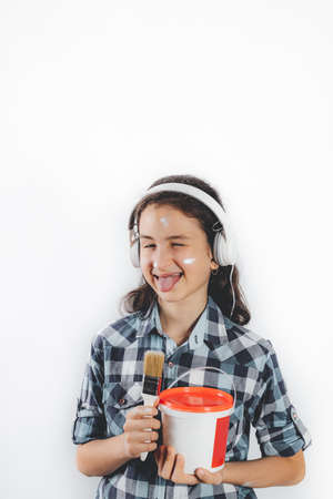 Portrait of a positive smiling teenage girl. Cute pretty girl in white headphones with a paint brush and a can of paint on a white background. positive emotion. vertical shotの写真素材