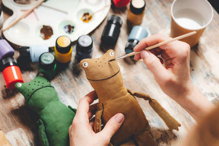 Handmade soft Doll frog. DIY gift crafting. Human hands hold frog soft toy. Needlework accessories on gray background. handmade house decor. Process of making a vintage soft toy. home workshop.の写真素材