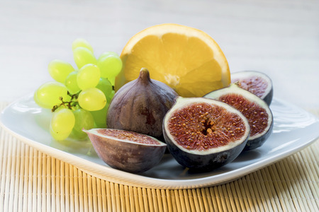 White plate with summer, juicy, sweet dietary, exotic fruits of figs, grapes, orange on a bamboo...