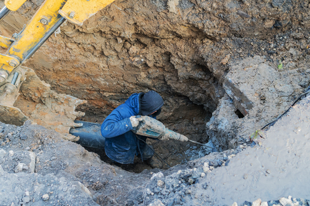 Workers excavate the pit with an excavator. Road repair. Pipeline failure. A man drills concrete with a puncher.の写真素材