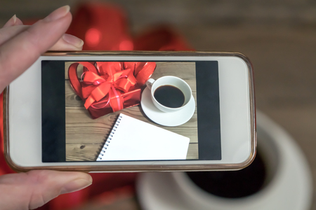 Red gift box with ribbon on a wooden background. A cup of coffee and a knitting heart. The whole picture is visible on the phone. Valentine's Day.の写真素材