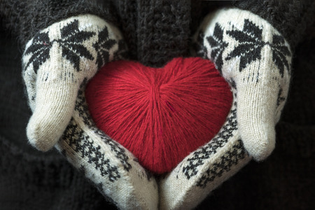 Hands are holding a red wool syrdze out of thread. Against a brown knitted sweater. Valentine's Day.