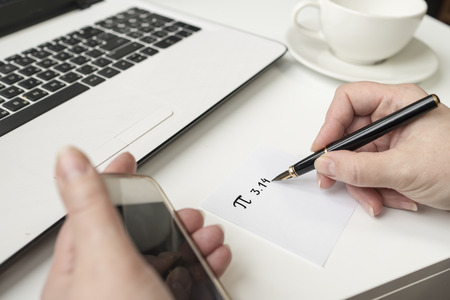 Happy Pi Day, written in black pen on white. On the desk a laptop, a cup, a hand holds the phone.の写真素材
