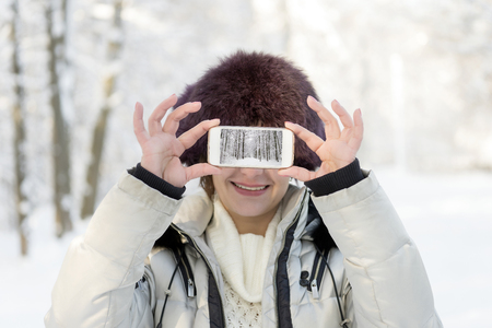 The girl in the fur hat is holding the phone at eye level. On the screen, the landscape is telephoned. Winter, day.の写真素材