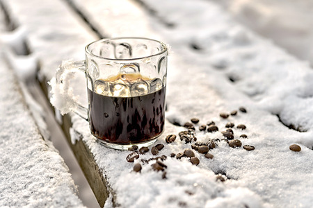 Mug of black espresso coffee stands in the snow, a wooden bench in the park, winter, scattered beansの写真素材