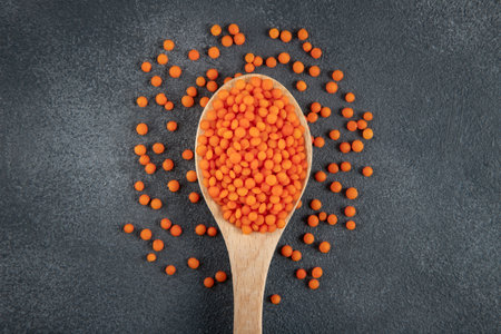 Red lentil grains in a wooden spoon on a gray background. Top view of dried orange lentils in a...