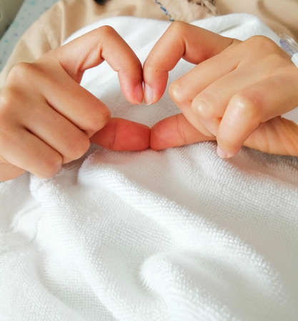 The patient who made a heart with her hands is sitting on the bed at the hospital with a saline solution line on hand.の写真素材