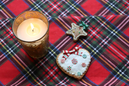 photo Burning candle in a glass candlestick and Christmas cookies on a red checkered tablecloth, top viewの写真素材