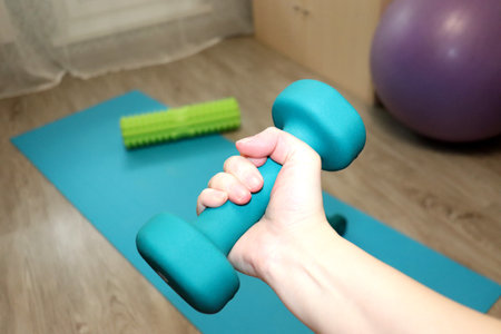 Blue dumbbell in a woman's hand against a fitness mat, fitball and roller in the room. Exercise for...