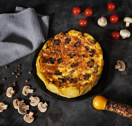 Homemade Lorraine pie with potatoes, chicken, mushrooms, cheese, onions and seasonings. In a black plate, on a dark table, cherry tomatoes, mushrooms, a gray napkin, microgreen and spices. の写真素材