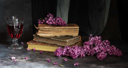 A stack of old books, a glass of red wine and a branch of beautiful fragrant lilacs on a dark background. Vintage still lifeの写真素材
