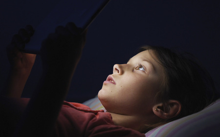 Caucasian girl lying in bed playing tablet in social internet in dark blue light under blacket. Childhood, communication and technology conceptの写真素材