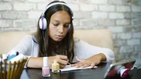 Cute teenager girl in headphones listening music on phone while making manicure applying nail polish with free copy space. Beauty, bodycare, wellness, hygiene, communication, leisure time concept.の写真素材