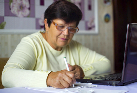 Senior old woman in eyeglasses checking costs of daily expenses on laptop at home. White caucasian female pensioner using computer calculating tax. Retirement, pension, technology, finance conceptの写真素材