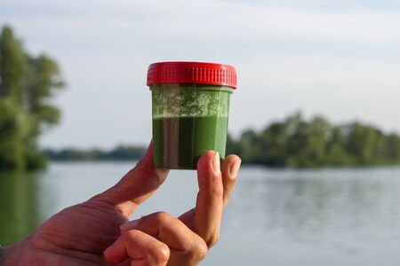 closeup fingers holding green algae sample with blurred river on background