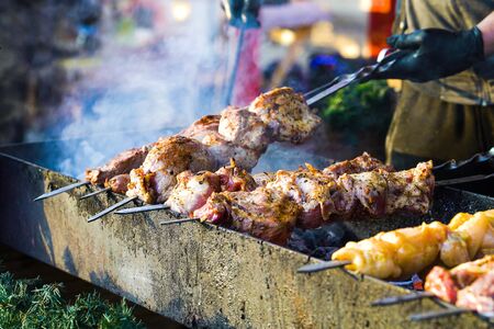 Juicy meat roasting on charcoal grill at holiday market stand decorated with spruce branches, smoke on background, selective focus. Crop male cook in gloves holding skewers. Concept of foodの写真素材
