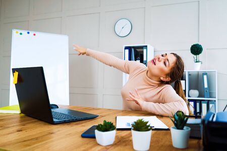 Selective focus on joyful female manager in casual wear sitting in office and dancing, laptop on table, flipchart and bookshelves on background. Portrait of young employee relaxing and having funの写真素材