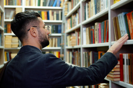 Male student with eyeglasses wearing jacket walking by bookshelves in search of literature,...