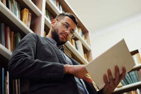 Young bearded man with eyeglasses standing next to bookshelves and reading book, flipping pages. Low...