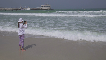 Teenage girl shoots on a smartphone the sea waves on Public Jumeirah Open Beach on the coast of the Persian Gulf, Dubaiの写真素材