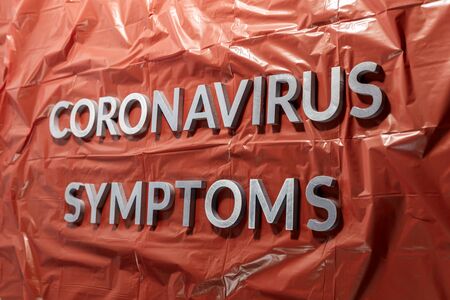 the words coronavirus symptoms laid with silver letters on crumpled orange plastic film - linear...