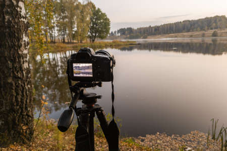 black digital camera on tripod shooting early foggy morning landscape at autumn lake with selective...