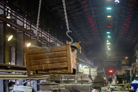Big rusty metal container hanging from chains, lifted by a crane inside the foundry. Close-up with...