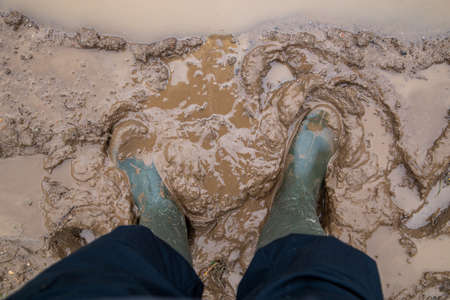 feet in green rubber boots stands in wet brown mud directly above view