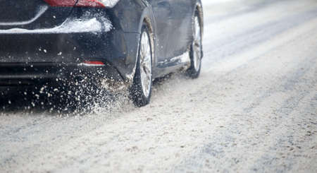 Road snow flinders flow from wheels of black car moving fast in daylight city with selective focus.の写真素材