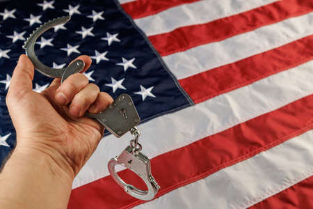caucasian hand holding silver metal handcuffs over US flag backgroundの写真素材