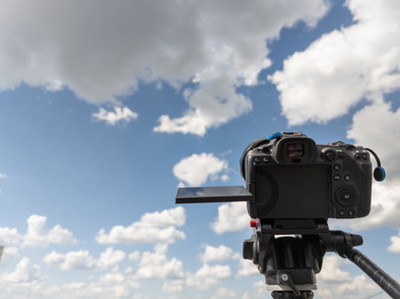 black professional digital camera with flip screen on a tripod pointed at blue sky with white clouds...