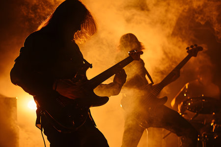 Long-haired rock musician with electric guitar playing in dark smoke with orange lighting.の素材
