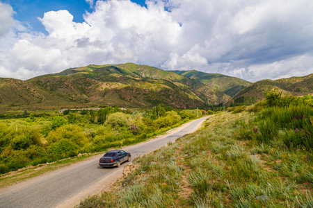 Car driving on country road with mountains, grass, and clouds in the backgroundの写真素材