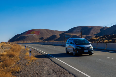 Car is driving along the road through natural mountain landscape with the flag of Kyrgyzstan is drawn on a mountain at sunny day.のeditorial素材