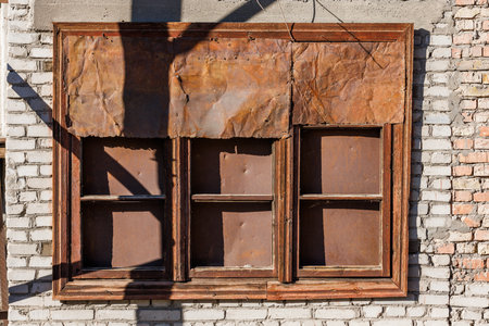 old brown wooden window covered with rusted sheet metal on white brick wall under direct sun lightの写真素材