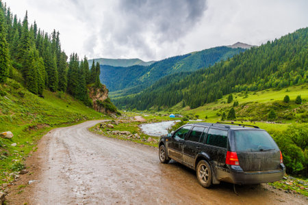 black car on a side of wet dirt road at green grass and forest covered mountain slopes at rainy...