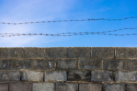 A low-angle shot of a gray concrete block wall topped with two strands of rusty barbed wire against a clear blue sky.の写真素材