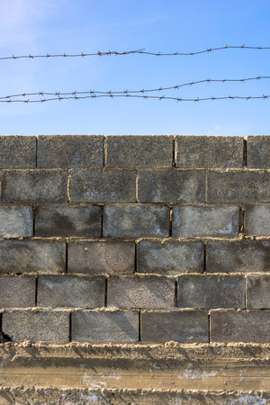 A low-angle shot of a gray concrete block wall topped with two strands of rusty barbed wire against a clear blue sky.の写真素材