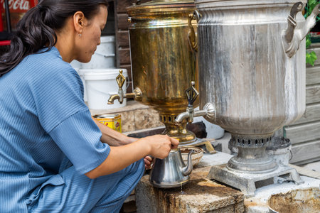 Cholok, Kyrgyzstan - July 16, 2024 - Focused woman making tea outdoors with samovar and teapotのeditorial素材
